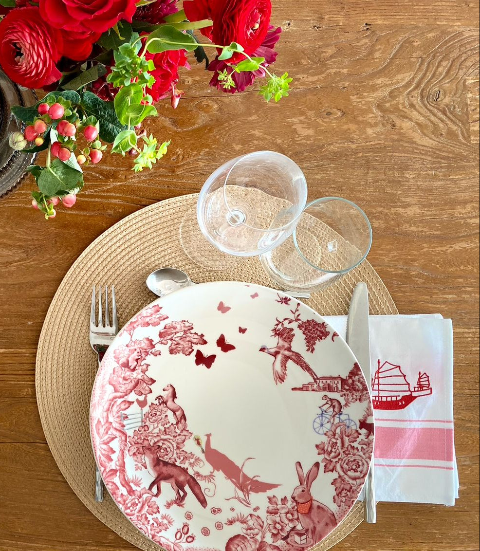 Dining table setting with red and white plates, silverware, cotton napkin with red junk embroidery.